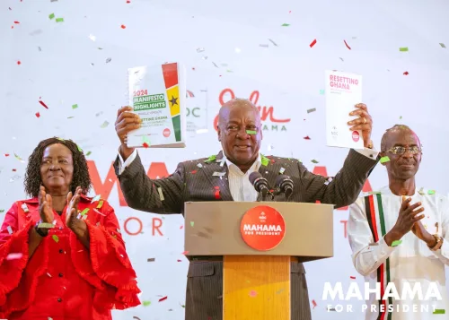 Presidential candidate of the NDC proudly holds up the 2024 manifesto above his head while displaying it to a lively audience. Standing to his left is the vice-presidential candidate, smiling confidently, and to his right is the NDC chairman, clapping in support. The trio is positioned against a backdrop of NDC party colors, emphasizing unity and readiness for the upcoming elections.