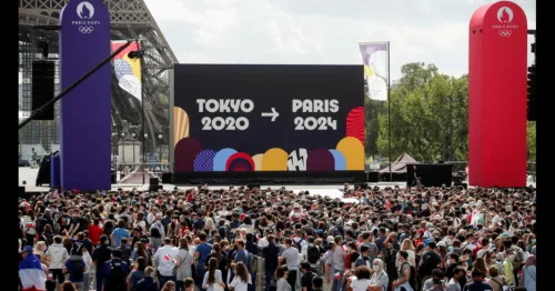 "Photo of the Paris 2024 Fan Zone, showcasing a large, bustling area with enthusiastic crowds. The scene includes multiple large screens for live event broadcasts, various food and merchandise stalls, and a stage where live music performances are taking place. The atmosphere is vibrant and festive, with flags and Olympic-themed decorations.