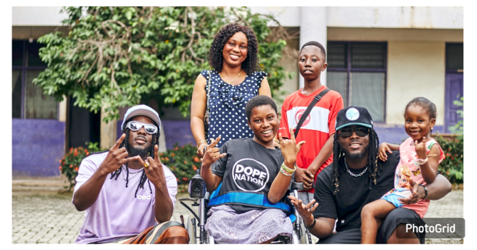 DopeNation, the Ghanaian music duo, stands beside Priscilla, a student from Serwaa Nyarko Senior High School, smiling together as they present her with an electronic wheelchair. Priscilla, visibly emotional and excited, holds the wheelchair with gratitude, while her mother looks on with pride and appreciation